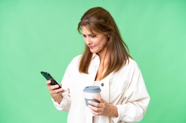 Orta yaşlı bir kadın izole edilmiş arka planda kahve ve bir cep telefonu tutuyor.