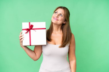Orta yaşlı beyaz bir kadın izole edilmiş arka planda bir hediye tutuyor gülümserken yukarı bakıyor.