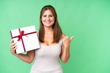 Orta yaşlı beyaz bir kadın, izole edilmiş arka plan üzerinde bir hediye tutuyor ve bir ürün sunmak için bir tarafa bakıyor.