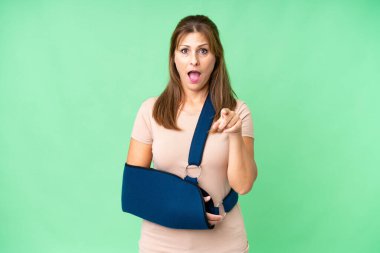 Orta yaşlı, kırık kollu, izole edilmiş arka planda askı takan şaşırmış ve ön tarafı işaret eden biri.