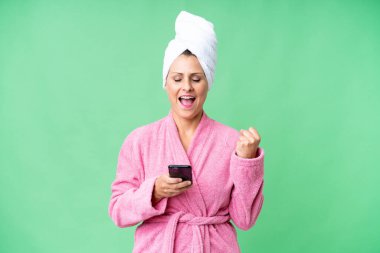 Bornozlu orta yaşlı beyaz kadın izole bir arka planda ve telefon zafer pozisyonunda.