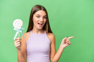 Kafkasyalı genç bir kız izole bir arka planda lolipop tutuyor. Şaşırmış ve parmağıyla kenarı işaret ediyor.