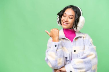 Arjantinli genç bir kadın izole edilmiş arka planda kışlık manşetler takıyor ve bir ürün sunmak için kenarı işaret ediyor.