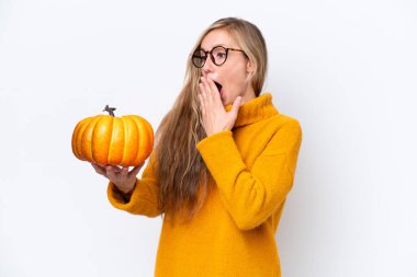 Beyaz arka planda izole edilmiş bir balkabağı tutan genç sarışın kadın şaşırmış ve şok olmuş bir yüz ifadesiyle