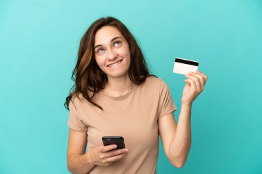 Kafkasyalı genç bir kadın mavi arka planda izole edilmiş. Cep telefonuyla kredi kartıyla alışveriş yapıyor.