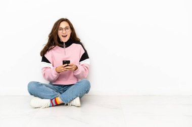 Beyaz arka planda izole bir şekilde yerde oturan beyaz bir kadın şaşırdı ve bir mesaj gönderiyor.