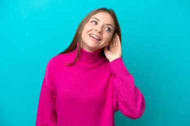 Genç Litvanyalı bir kadın mavi arka planda izole edilmiş bir şekilde elini kulağına koyarak bir şeyler dinliyor.