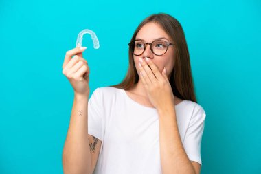 Görünmez diş telleri tutan genç Litvanyalı kadın mavi arka planda şaşırmış ve şok olmuş bir yüz ifadesiyle izole edilmiş.