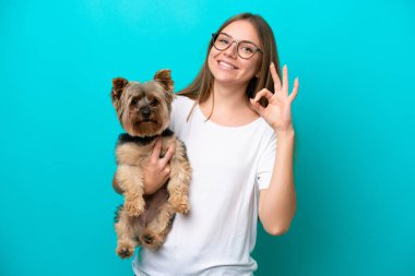 Mavi arka planda köpeği tutan Litvanyalı genç bir kadın parmaklarıyla tamam işareti gösteriyor.