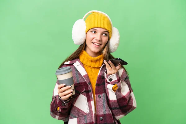 年轻的高加索女孩穿着冬衣 背景偏僻 手里拿着咖啡和手机 — 图库照片