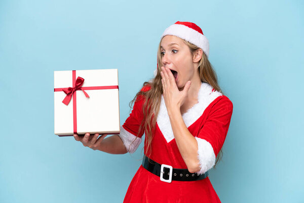 Young blonde woman dressed as Mama claus holding a present isolated on blue background with surprise and shocked facial expression