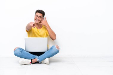 Young man sitting on the floor isolated on white background making phone gesture and pointing front