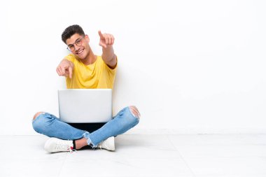 Genç bir adam beyaz arka planda izole bir şekilde yerde oturuyor. Gülümserken seni işaret ediyor.