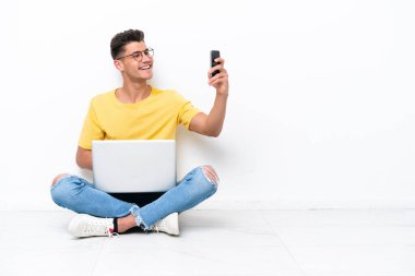 Yerde oturan genç adam beyaz arka planda izole bir şekilde selfie çekiyor.