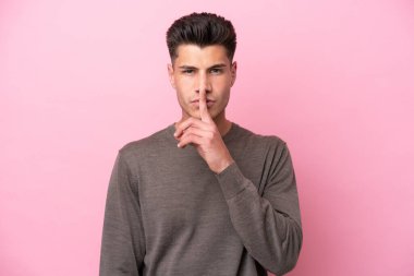 Pembe arka planda izole edilmiş genç beyaz adam sessiz bir jest işareti gösteriyor. Parmağını ağzına sokuyor.