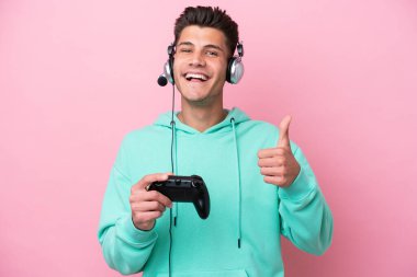 Pembe arka planda izole edilmiş bir bilgisayar oyunu kontrolörüyle oynayan yakışıklı beyaz bir adam baş parmak işareti yapıyor.