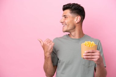 Elinde pembe arka planda izole edilmiş patates kızartması tutan genç beyaz bir adam bir ürünü sunmak için kenarı işaret ediyor.