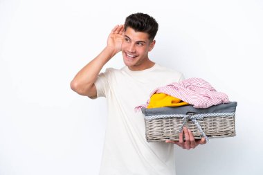 Beyaz arka planda çamaşır sepetini tutan genç beyaz bir adam elini kulağına koyarak bir şeyler dinliyor.