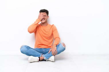 Genç adam, beyaz arka planda izole bir şekilde yerde oturuyor. Gözleri elleriyle kaplıyor ve gülümsüyor.