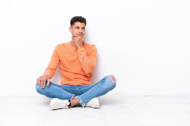 Beyaz arka planda izole bir şekilde yerde oturan genç adamın şüpheleri ve kafası karışmış bir yüz ifadesi var.