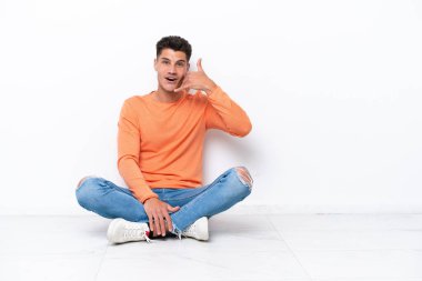 Yerde oturan genç adam beyaz arka planda izole bir şekilde telefon jesti yapıyor. Bana geri dönüş işareti ver.