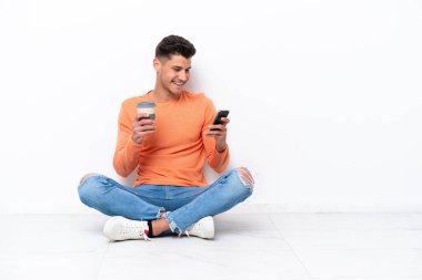 Genç adam beyaz arka planda izole bir şekilde yerde oturuyor. Elinde kahve ve cep telefonu var.
