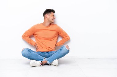 Yerde oturan genç adam, beyaz arka planda izole bir şekilde, çabaladığı için sırt ağrısından muzdarip.
