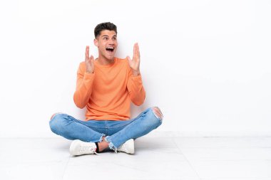 Yerde oturan genç adam, beyaz arka planda, şaşırmış bir yüz ifadesiyle izole edilmiş.