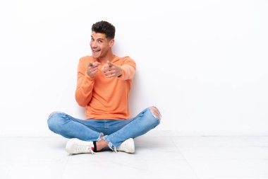 Genç adam beyaz arka planda izole bir şekilde yerde oturuyor. Önü işaret ediyor ve gülümsüyor.