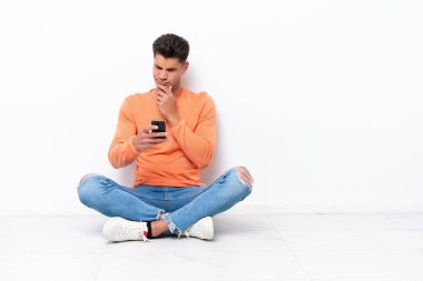 Genç adam beyaz arka planda izole bir şekilde yerde oturuyor. Düşünüyor ve bir mesaj gönderiyor.
