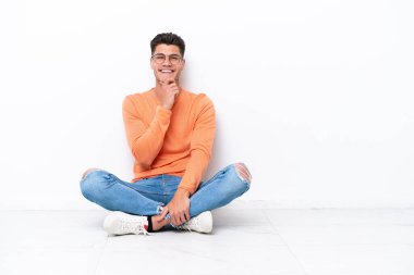 Yerde oturan genç adam beyaz arka planda gözlüklü ve gülümseyen biri.