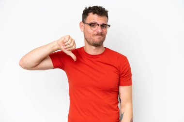 Beyaz arka planda izole edilmiş beyaz tenli yakışıklı genç adam olumsuz ifadeyle baş parmağını aşağı gösteriyor.