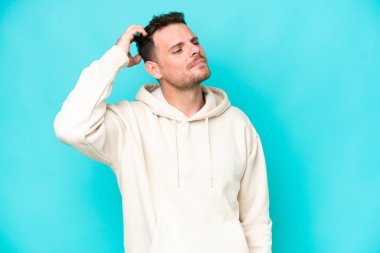 Kafkasyalı yakışıklı genç adam mavi arka planda izole edilmiş, kafası kaşınırken şüpheleri var.