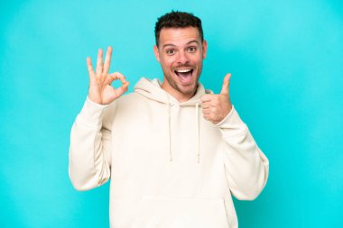 Mavi arka planda izole edilmiş beyaz, yakışıklı genç bir adam tamam işareti ve baş parmak hareketi gösteriyor.