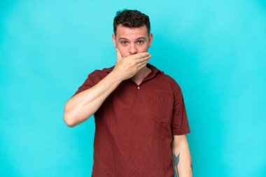Beyaz tenli yakışıklı bir adam mavi arka planda izole edilmiş. Ağzını eliyle kapatıyor.