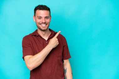 Mavi arka planda izole edilmiş beyaz, yakışıklı genç bir adam bir ürün sunmak için kenarı işaret ediyor.