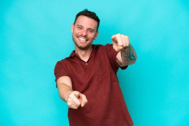 Genç beyaz yakışıklı adam mavi arka planda izole edilmiş mutlu bir ifadeyle ön tarafı işaret ediyor.