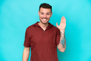 Beyaz tenli yakışıklı adam mavi arka planda izole edilmiş mutlu bir ifadeyle selamlıyor.