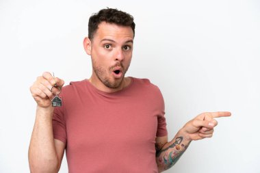 Beyaz arka planda izole edilmiş, şaşırmış ve işaret eden ev anahtarlarını elinde tutan genç beyaz bir adam.