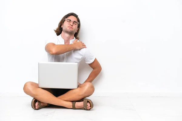 Young handsome man with a laptop sitting on the floor isolated on white background suffering from pain in shoulder for having made an effort