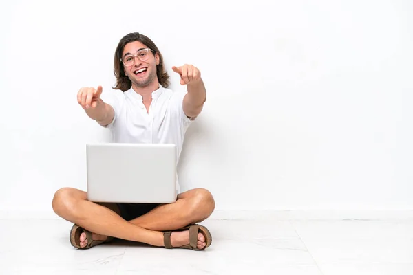 Young handsome man with a laptop sitting on the floor isolated on white background points finger at you while smiling
