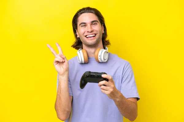 若いですハンサムなCaucasian男遊びますとともにAビデオゲームコントローラ上の隔離された上に黄色の背景笑顔と勝利サインを示します — ストック写真