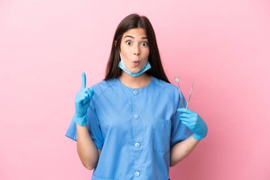 Pembe arka planda izole edilmiş aletleri tutan dişçi kadın parmağını kaldırırken çözümü fark etmeyi amaçlıyor.