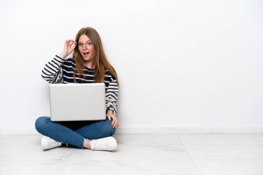 Young caucasian woman with a laptop sitting on the floor isolated on white background with glasses and surprised