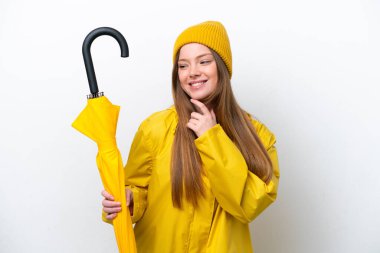 Beyaz arka planda izole edilmiş yağmurluk ve şemsiyeli beyaz kadın yan tarafa bakıyor ve gülümsüyor.