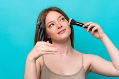 Mavi arka planda izole edilmiş beyaz bir kadın elinde makyaj fırçası tutuyor.