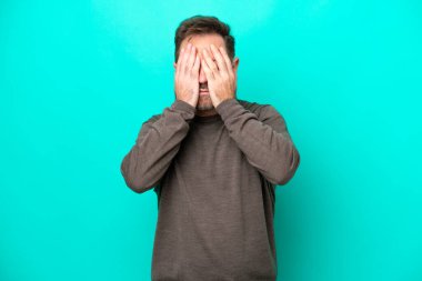 Orta yaşlı, beyaz bir adam mavi arka planda izole edilmiş, gözleri elle kapatılmış.