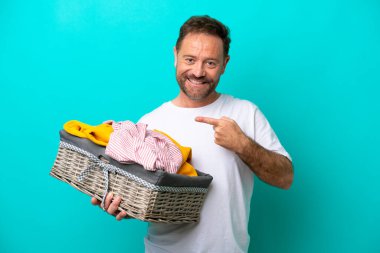 Orta yaşlı bir kadın mavi arka planda izole edilmiş bir çamaşır sepetini elinde tutuyor. Ürünü sunmak için yan tarafa bakıyor.