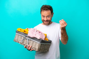 Mavi arka planda izole edilmiş çamaşır sepeti tutan orta yaşlı bir kadın para hareketi yapıyor.