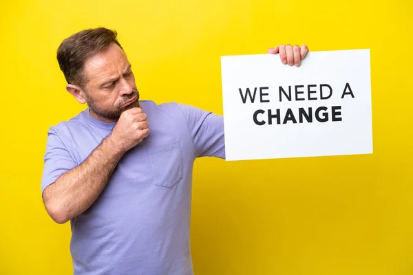 stock image Middle age caucasian man isolated on yellow background holding a placard with text We Need a Change and thinking